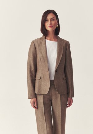 Vrouw draagt een bruine, op maat gemaakte blazer en bijpassende broek over een wit overhemd, staande tegen een effen lichte achtergrond.
