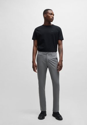 T-shirt noire à manches courtes avec logo ; pantalon slim gris clair ; tissu lisse ; baskets noires ; modèle posé sur un fond uni.