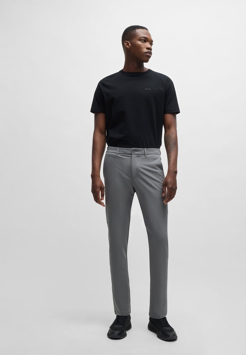 Černé tričko s krátkým rukávem a logem; světle šedé slim-fit kalhoty; hladká látka; černé tenisky; model stojí před jednobarevným pozadím.