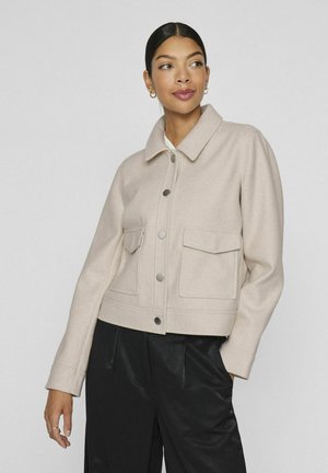Chaqueta corta beige hecha de un tejido suave, con un cierre de botones en el frente, dos bolsillos de parche y un cuello clásico. Combinada con pantalones negros.