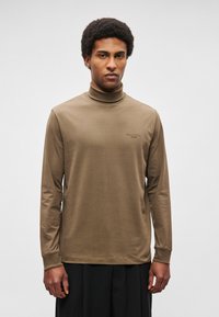 Pikad varrukad, kõrge kaelusega särk oliivrohelisest puuvillast, millel on rinnal peen logo ja keha kujule sobivad varrukad sujuva silueti saavutamiseks.