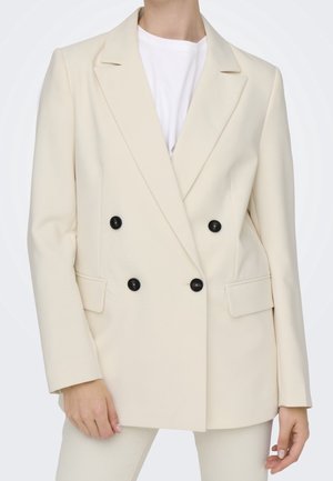 Femme portant un blazer croisé crème avec des boutons noirs sur une chemise blanche, debout devant un fond uni.