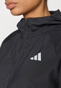 Veste noire à capuche avec une fermeture éclair intégrale et un tissu texturé. Logo réfléchissant distinctif avec trois bandes sur le côté droit de la poitrine.