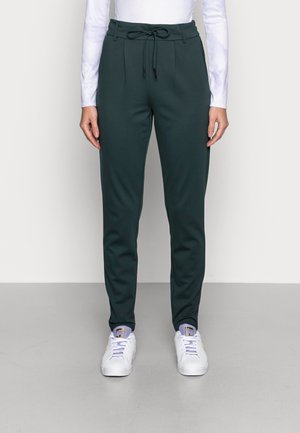 Pantalon de jogging vert foncé avec taille à cordon de serrage, jambes fuselées et texture lisse, associé à des baskets blanches avec des accents lilas.