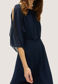 Mörkblå klänning med genomskinliga, flödande ärmar och elastisk midja. Slät tyg, samlad halslinning och utskärningar vid axlarna.