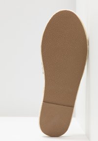 Semelle d'une espadrille beige avec un dessous en caoutchouc texturé, présentée verticalement sur un fond blanc.