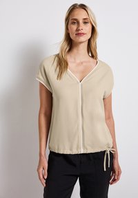 Beige bluse med korte ærmer, V-udskæring og snor i kanten. Har subtile hvide accenter og en glat tekstur.