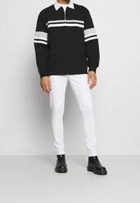 Black long-sleeve shirt with zip neck, white collar, and gray/white stripes on sleeves, paired with white slim-fit pants and black boots.
