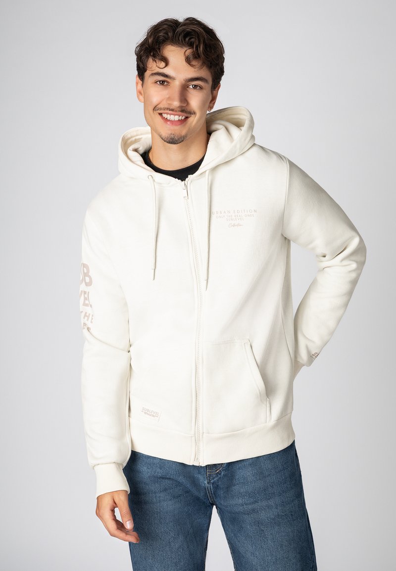 Cremefarbene Zip-Hoodie mit Kapuze, Fronttaschen und Textdesign auf der linken Brust und dem Ärmel. Aus weichem Baumwollmaterial.