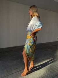 A woman with long blonde hair stands in profile, wearing a light blue top and a colorful, patterned skirt, against a neutral background.