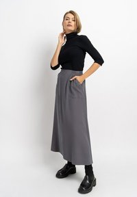 Schwarzer Rollkragenpullover kombiniert mit einem langen, grauen, elastischen Rock mit zwei vorderen Taschen. Das Model trägt schwarze Schuhe mit dicken Sohlen.