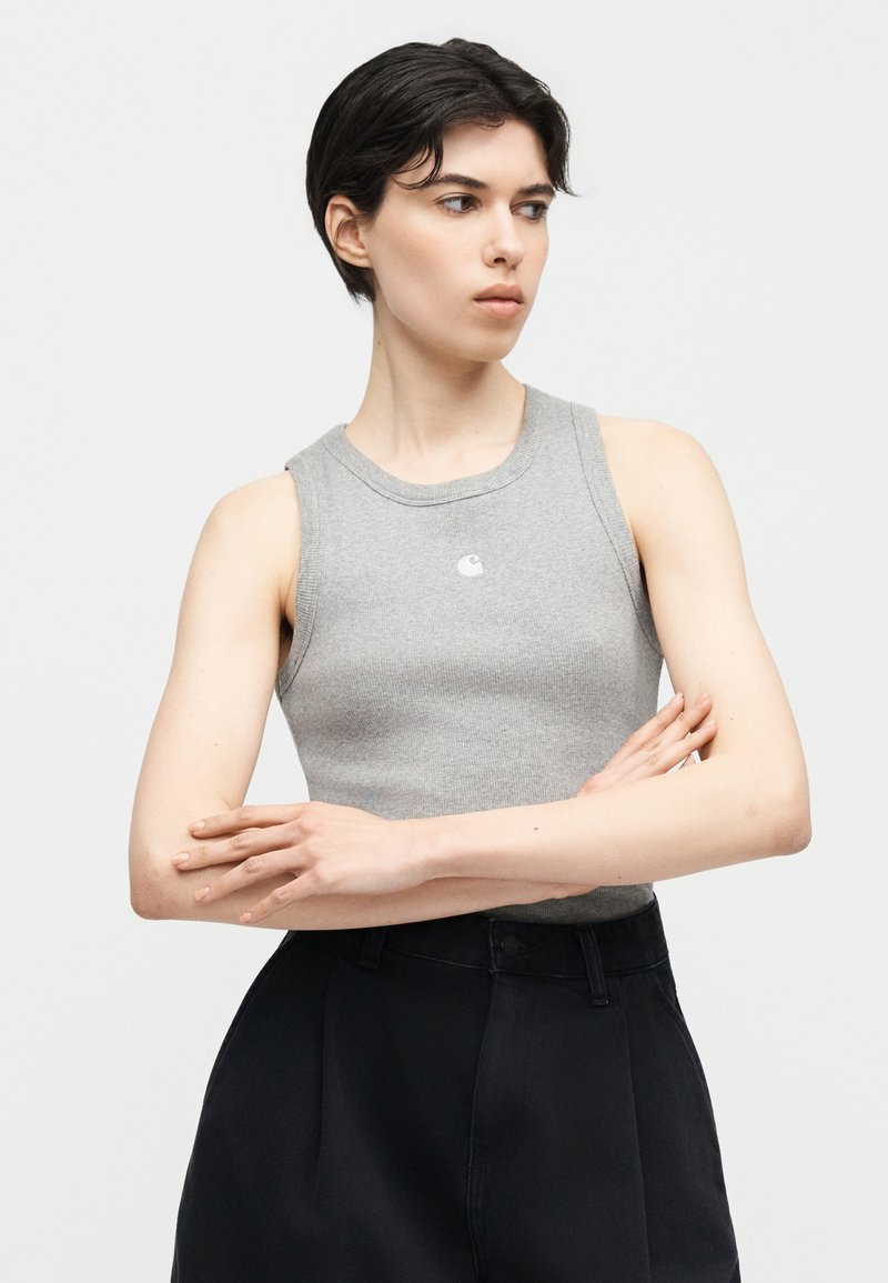 Personne aux cheveux courts et foncés, portant un haut gris côtelé sans manches et un pantalon noir taille haute, les bras croisés, regardant sur le côté.