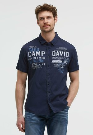 Mann trägt ein marineblaues Kurzarmhemd mit weißem und blauem Druck, kombiniert mit blauen Jeans.