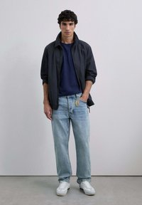 Un joven se encuentra de pie con una mano en el bolsillo, vistiendo una chaqueta negra, una camisa azul marino, jeans claros con llavero y zapatillas blancas.
