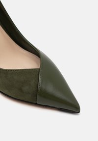 Zapato de tacón alto verde con punta afilada, que combina cuero suave y ante texturizado con un diseño de corte.