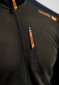 Mørkegrønt fleecejakke med blå accenter, der har en fremtrædende frontlynlås og en orange logo-tag. Glat tekstur med lynlåslomme.