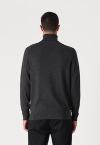 Ein Mann trägt einen dunkelgrauen Rollkragenpullover und schwarze Hosen, von hinten auf einem schlichten weißen Hintergrund abgebildet.