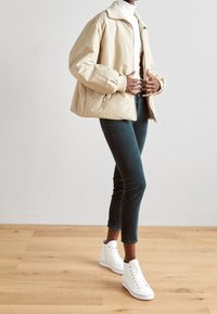 Beige puffjakke med høj krave, lynlåslommer og en afslappet pasform, parret med mørkegrønne jeans og hvide højtoppede sneakers.