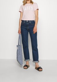 Ljusrosa T-shirt med "LEVI'S" logotyp, mörkblå högmidjade jeans, svarta sandaler och en randig tote-väska. Avslappnad outfit med en lös passform.