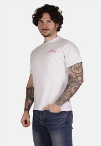 T-shirt bianca in cotone con collo rotondo e maniche corte, caratterizzata da un piccolo logo ricamato rosa sul lato sinistro del petto.