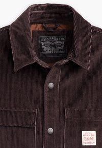 Brown corduroy shirt with a textured finish, featuring a button-up front, collar, and patch with brand details on the left pocket.