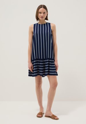 Frau steht und trägt ein ärmelloses dunkelblaues Kleid mit vertikalen Streifen am Oberteil und horizontalen Streifen am gerafften Rock, kombiniert mit braunen Slide-Sandalen.