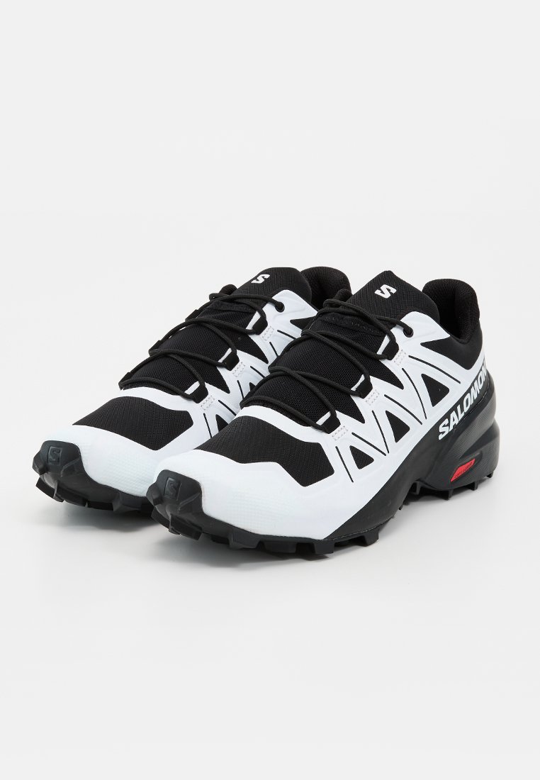 Paire de chaussures de trail running Salomon noires et blanches avec semelles épaisses et lacets noirs sur fond blanc uni.
