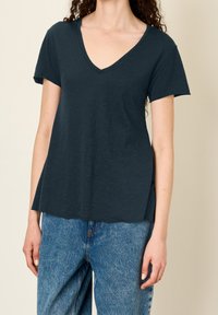 Žena v tmavě modrém topu s výstřihem do V a krátkým rukávem, který je zkombinovaný se světle modrými volnými džíny s vysokým pasem, na béžovém pozadí.