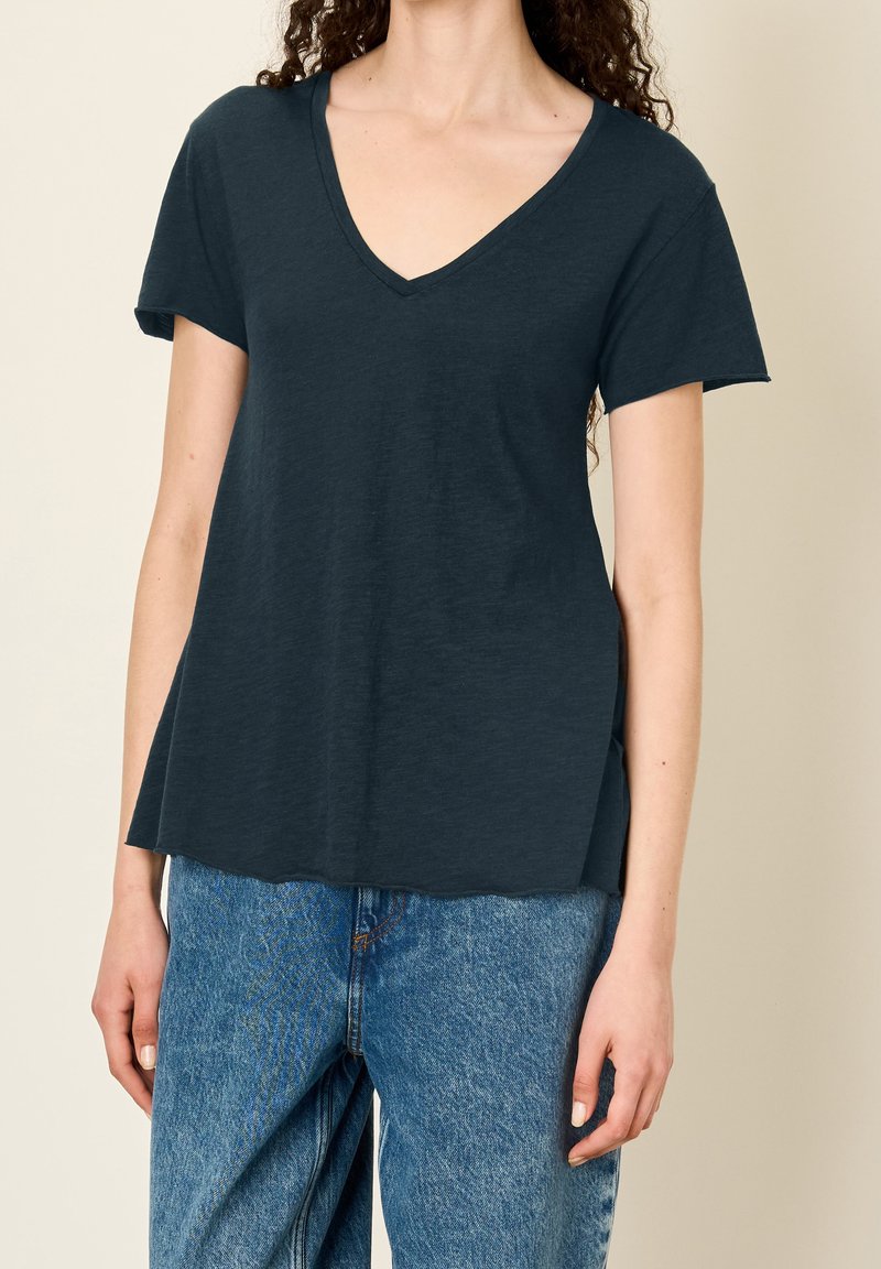 Žena v tmavě modrém topu s výstřihem do V a krátkým rukávem, který je zkombinovaný se světle modrými volnými džíny s vysokým pasem, na béžovém pozadí.