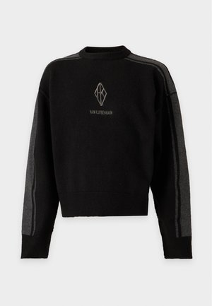 Mann, der einen schwarzen Pullover mit grauen Streifen und Logo über einem beige karierten Hemd trägt, schwarze weite Hosen und schwarze Schuhe, steht mit einer Hand in der Hosentasche.