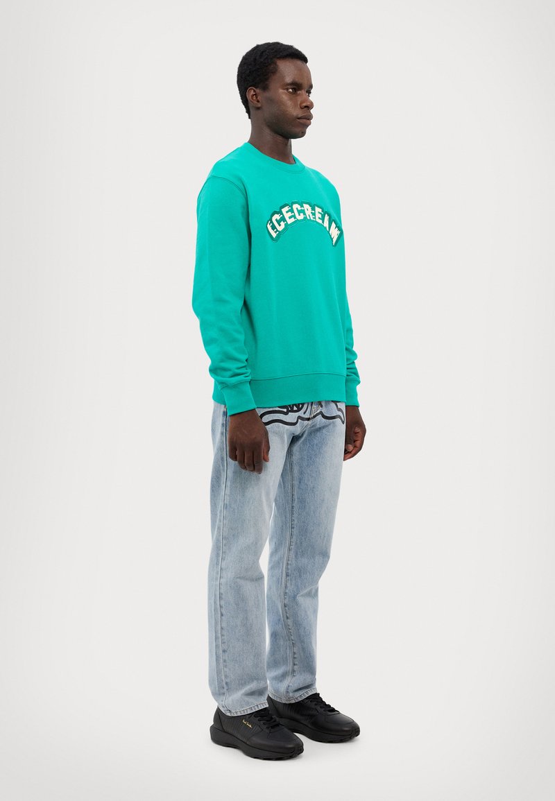 Felpa a girocollo turchese con logo "ICECREAM", abbinata a jeans azzurri chiari e sneakers nere, in piedi contro uno sfondo neutro.