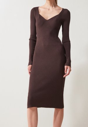 Femme portant une robe midi ajustée, côtelée, à manches longues, de couleur brun foncé avec un large décolleté en V, sur un fond uni.