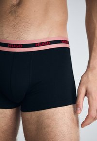 Boxeri scurți negri din bumbac cu un brăc de culoare roz, având inscripționat în roșu "HUGO". Textură netedă și design ajustat. Mână odihnită pe coapsă.