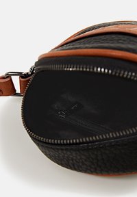 Pyöreä laukku, joka on valmistettu teksturoidusta mustasta nahasta ja jossa on ruskea nahkainen reunus. Sisäpuolella on vetoketjutasku ja sileä musta vuori.