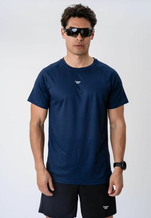 Mand iført sorte sports solbriller, navyblå kortærmet skjorte og sorte shorts, der står mod en ensfarvet lys baggrund.