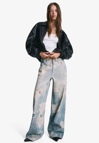 Schwarze Lederjacke im Oversized-Stil, weißes enges Tanktop und hellblaue weit geschnittene Jeans mit floralen Mustern und kleinen Akzentfarben.