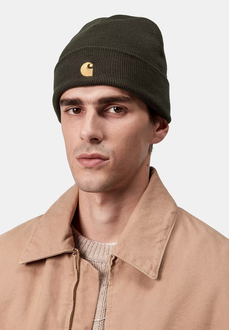 Gorro de punto verde oscuro con un detalle del logo en dorado. Lo lleva una persona con una chaqueta beige, mostrando una textura acanalada y un dobladillo plegable.