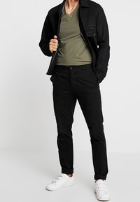 Czarna dżinsowa kurtka noszona na oliwkowej koszulce z dekoltem w kształcie V, w zestawie z czarnymi spodniami i białymi trampkami. Prosty design, dopasowany krój.