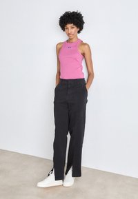 Top senza maniche rosa con logo, pantaloni neri larghi e scarpe da ginnastica bianche. Texture liscia, design casual, leggere aperture laterali nei pantaloni.