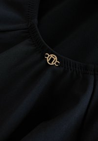 Crna tkanina s mekom teksturom sadrži nabubrene detalje i metalni logo naglašen sprijeda, okružen prozračnim mrežastim materijalom.