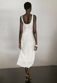 Weißes ärmelloses Kleid mit tiefem Rückenausschnitt, das aufwendige Spitzenverzierungen am Saum aufweist. Glatter Stoff und eine lockere Passform. Einfaches weißes Armband.