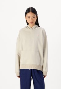 Felpa beige con cappuccio, con orlo e polsini a costine, dotata di tasca a marsupio frontale e un piccolo patch con logo sul petto.