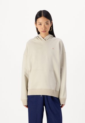 Felpa beige con cappuccio, con orlo e polsini a costine, dotata di tasca a marsupio frontale e un piccolo patch con logo sul petto.
