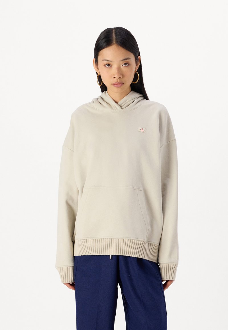 Felpa beige con cappuccio, con orlo e polsini a costine, dotata di tasca a marsupio frontale e un piccolo patch con logo sul petto.