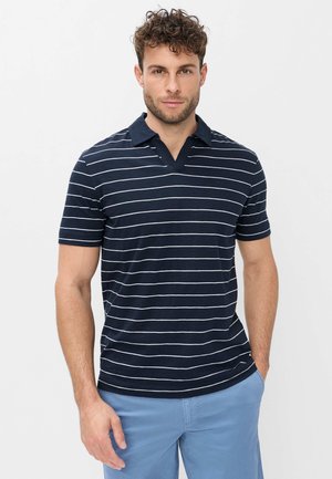 Mand med krøllet hår iført navyblå og hvide stribede polo og lyseblå shorts, stående med den ene hånd i lommen.