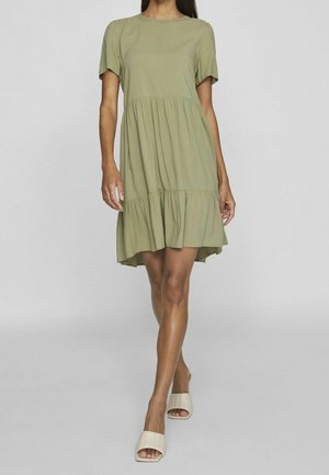 Frau trägt ein kurzärmeliges, hellolivgrünes Stufenkleid, kombiniert mit beigen offenen Sandalen, steht vor einem einfarbigen Hintergrund.