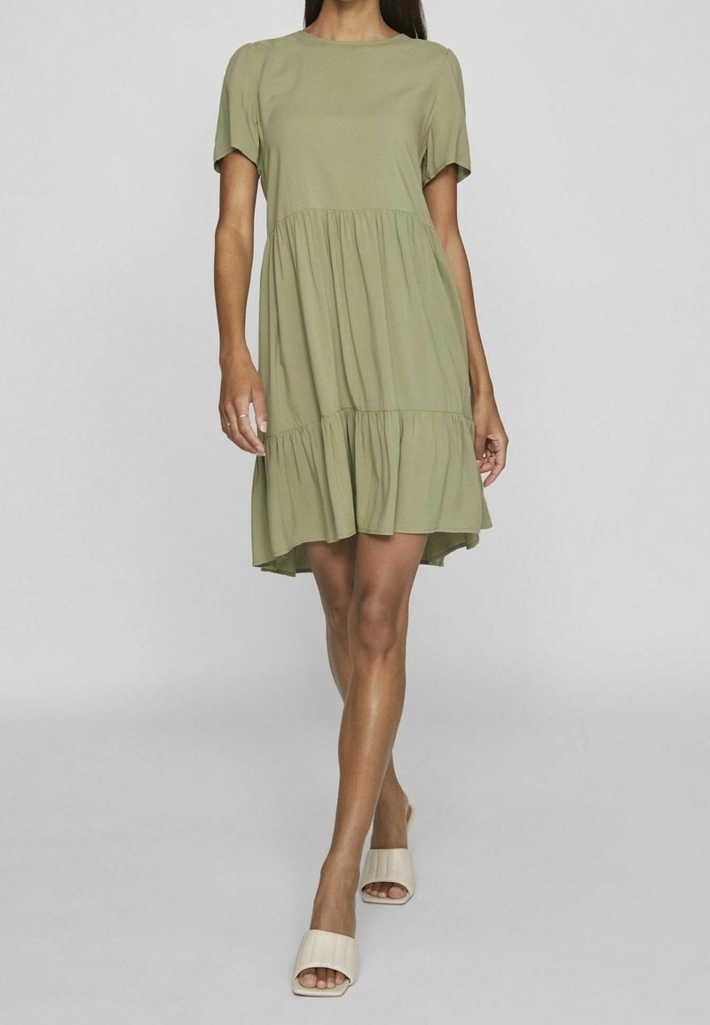 Femme portant une robe courte à volants manches courtes de couleur vert olive clair, associée à des sandales ouvertes beiges à enfiler, debout devant un fond uni.