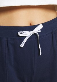 Marineblauwe sweatpants met een brede tailleband en witte trekkoord met "TAILOR" erop gedrukt. Gladde textuur, minimalistisch ontwerp, zonder zakken.