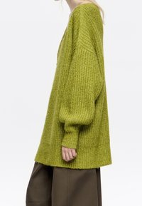 Personne portant un pull ample en grosse maille vert citron avec des manches ballon et un pantalon large marron foncé, vue de profil.