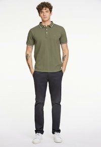 Olivengrøn polo-shirt med tre knapper og krave, prydet med stribede detaljer. Kombineret med mørke bukser og hvide sneakers.